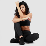 a fit woman setting on ground with pose and look at the camera ,she wearing matching legging and bra black with shoes same color ,plus he background is light grey 