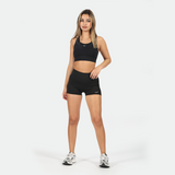 a fit woman wearing matching black set is  sports bra and short with logo ,and she wearing shoes ,plus the background is light grey 
