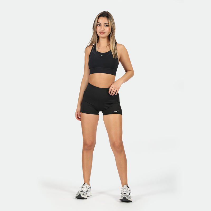 a fit woman wearing matching black set is  sports bra and short with logo ,and she wearing shoes ,plus the background is light grey 