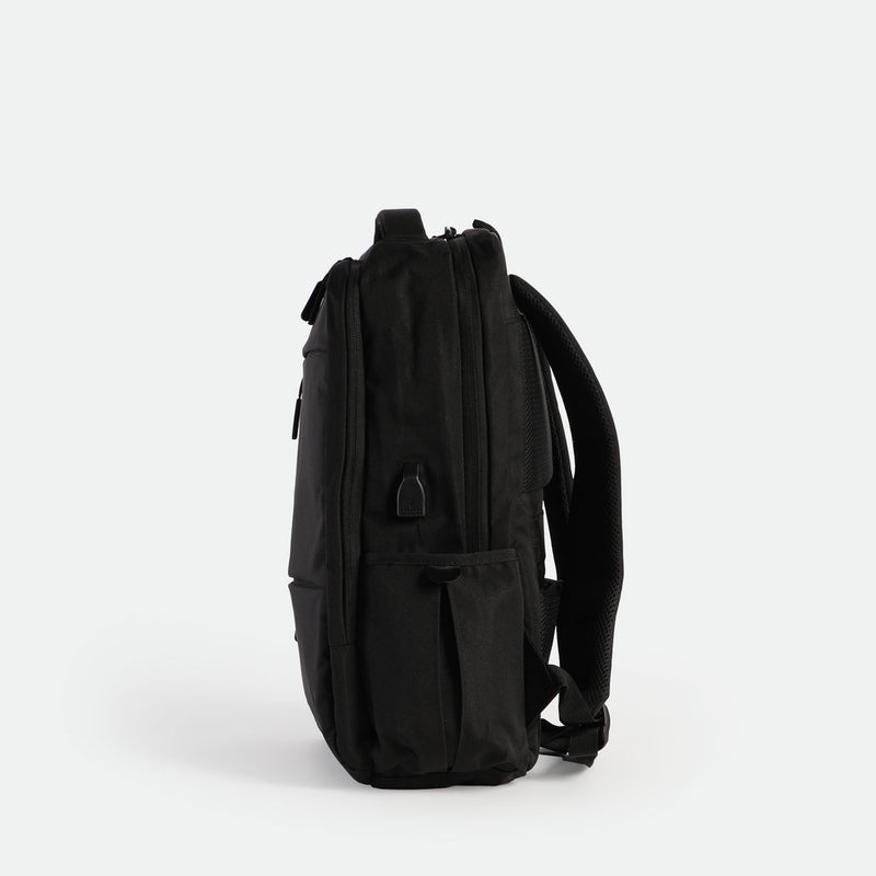 A sleek, black backpack is shown standing upright on its base, viewed from the side against a plain white background. The design is minimalist with subtle zippers and padded shoulder straps visible.