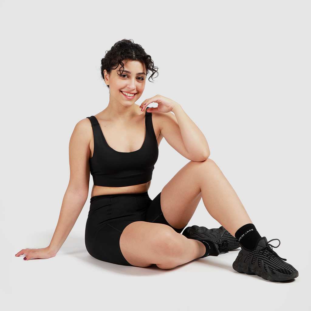 A woman wearing a matching bra and legging, sitting on the floor looking down, smiling, wearing black shoes