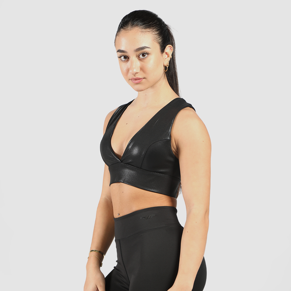 A fit woman with dark hair in a sleek ponytail wears a black faux-leather sports bra with thick straps and high-waisted leggings. She stands against a white background, gazing confidently at the camera.