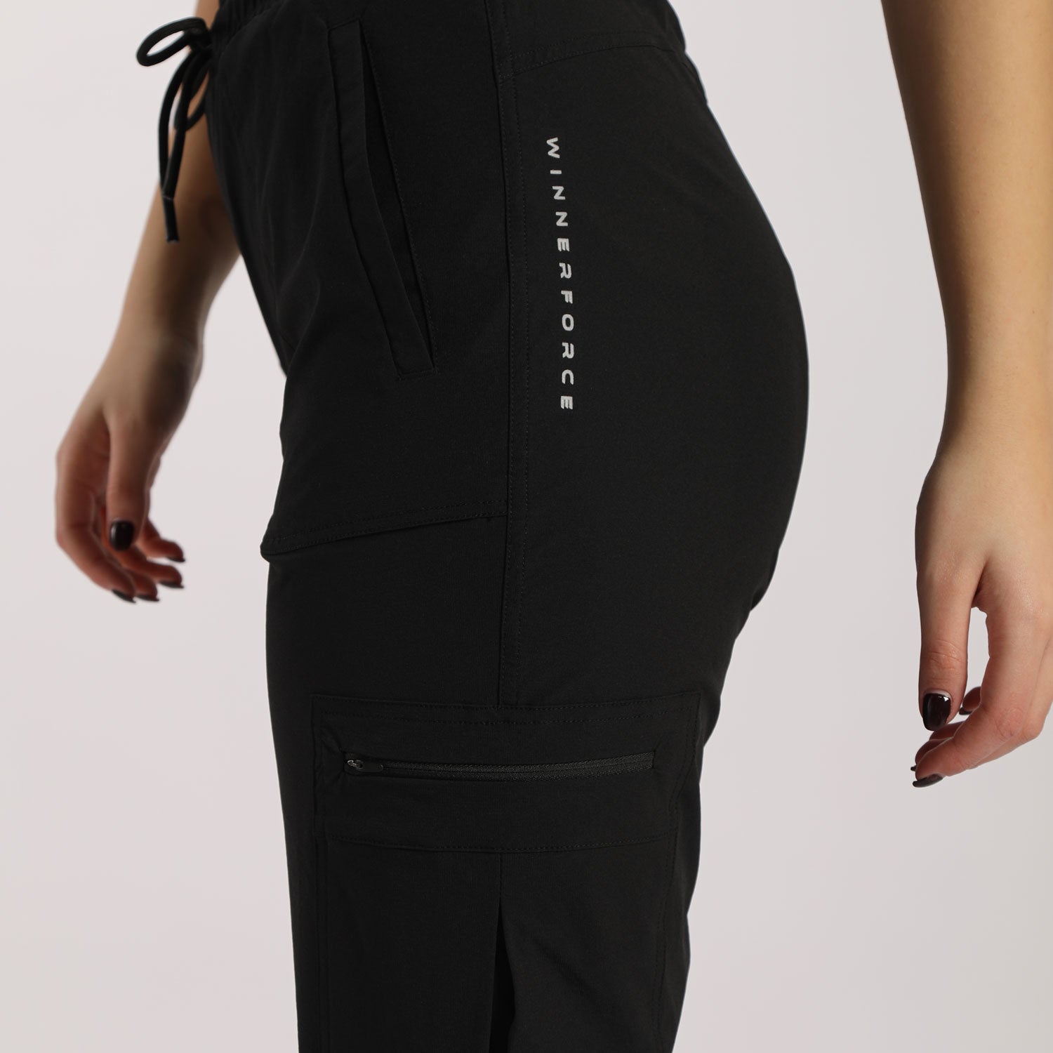 A close-up view of a person wearing black cargo jogger pants with multiple pockets, including a zippered pocket on the thigh and a vertical printed text design on the side. The pants have an elastic waistband with a drawstring