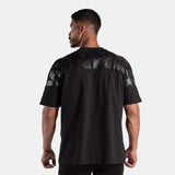 Man in black T-shirt with glossy print on back, facing away. Neutral gray background highlights the shirt's sleek design and casual tone.
