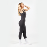 a fit woman stand at right and look at up , she wearing jump suit black with logo and socks white ,the background is light grey  