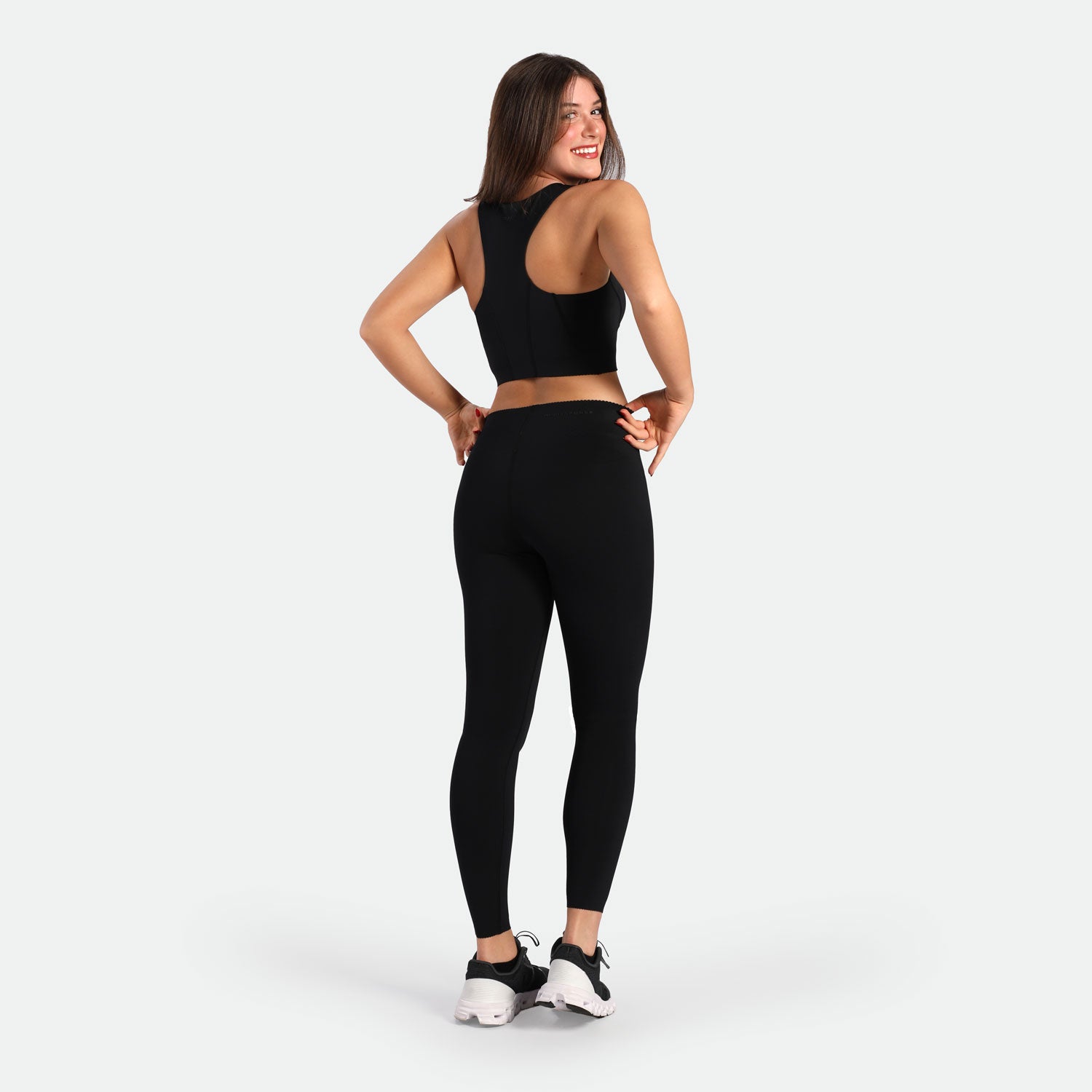 Smiling woman in black athletic wear and sneakers stands in a confident pose against a white background, conveying a sense of fitness and joy.