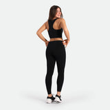 Smiling woman in black athletic wear and sneakers stands in a confident pose against a white background, conveying a sense of fitness and joy.