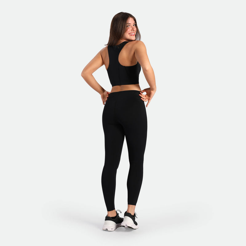 Smiling woman in black athletic wear and sneakers stands in a confident pose against a white background, conveying a sense of fitness and joy.