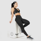 Woman posing, looking right, wearing a faux leather bra and leggings, plus a white carrier bearing the logo