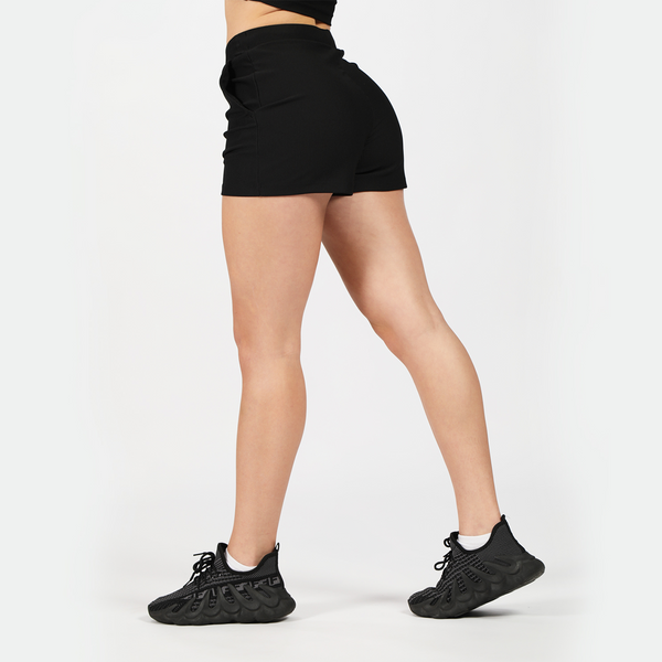 a fit woman stand at right and wearing short black with shoes ,plus the background is light grey 