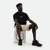 Man sitting on a box wearing a black t-shirt with "WINDFORCE" text, beige shorts, and black sneakers. He exudes a calm and relaxed vibe.