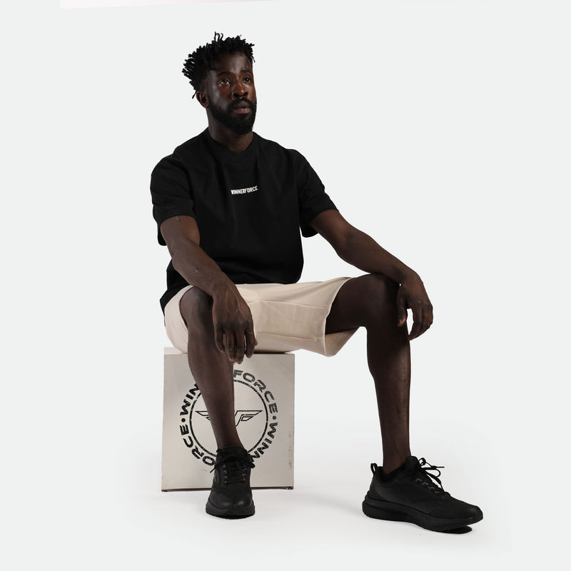 Man sitting on a box wearing a black t-shirt with "WINDFORCE" text, beige shorts, and black sneakers. He exudes a calm and relaxed vibe.