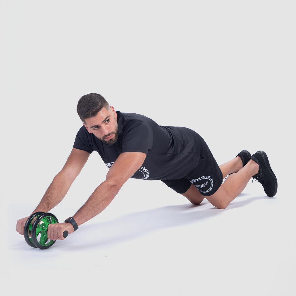 A man in a black athletic outfit is using an ab roller on his knees. He extends his arms forward, focused and concentrated, against a plain white background.