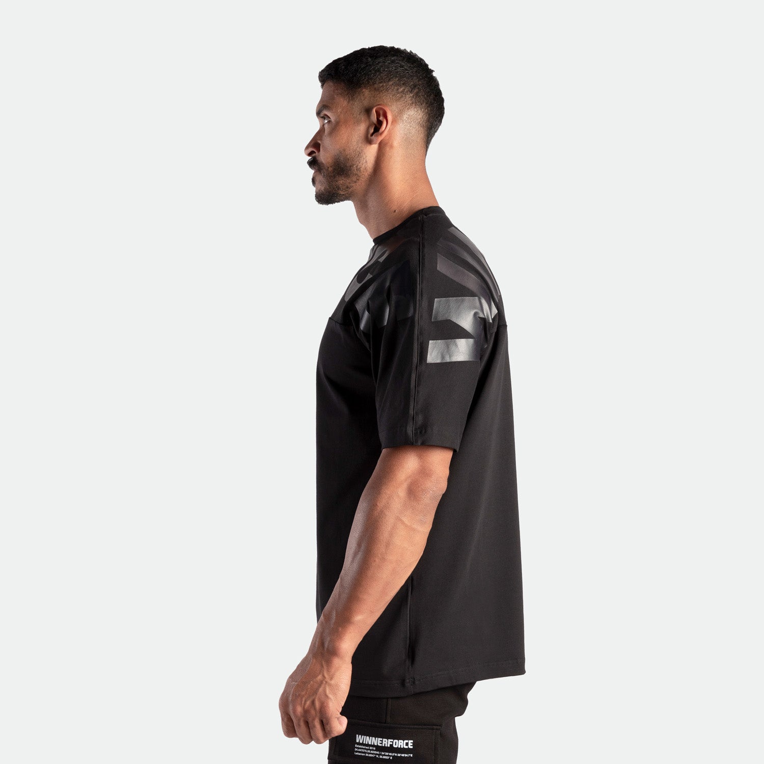 A man in a profile view wearing a black T-shirt with graphic details on the sleeves stands against a light gray background, conveying a sporty, confident look.