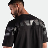 Man in black T-shirt with glossy print on back, facing away. Neutral gray background highlights the shirt's sleek design and casual tone.
