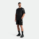 Man in sleek black sportswear stands confidently. He's wearing a sporty black t-shirt, shorts, socks, and sneakers against a plain white background.