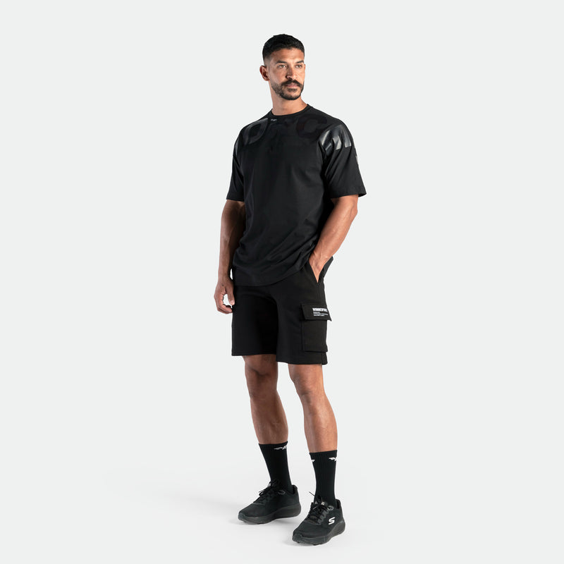 Man in sleek black sportswear stands confidently. He's wearing a sporty black t-shirt, shorts, socks, and sneakers against a plain white background.