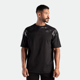 A man in a black T-shirt with shiny details looks to the right. He has short dark hair and a beard, exuding a confident and relaxed demeanor.