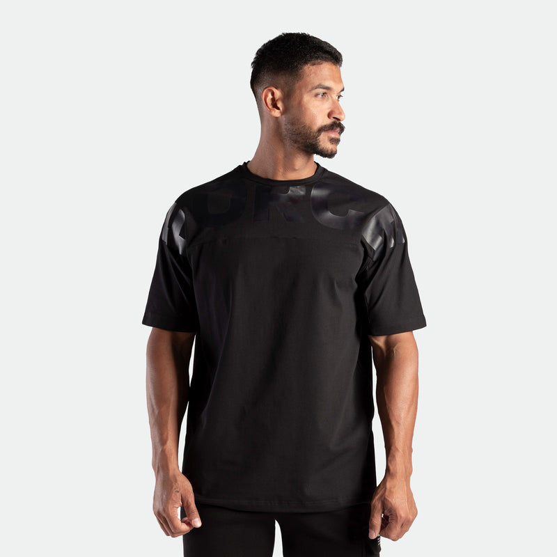 A man in a black T-shirt with shiny details looks to the right. He has short dark hair and a beard, exuding a confident and relaxed demeanor.