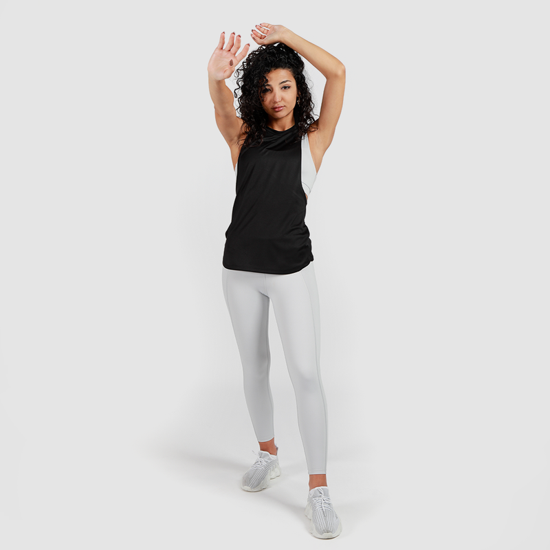 Woman with curly hair in a black tank top and white leggings stretches with arms overhead. She is wearing white sneakers on a plain background.