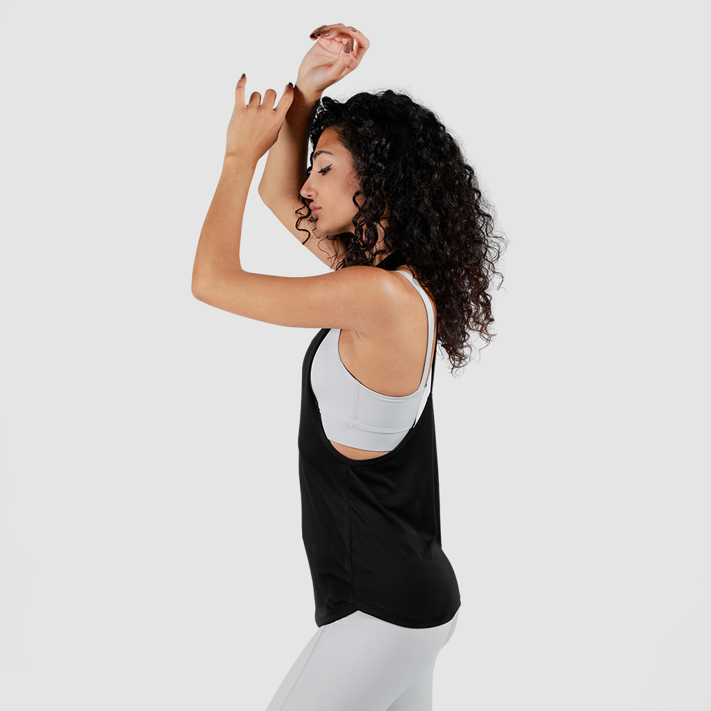 A woman with curly hair, in a white sports bra and black tank top, poses gracefully with raised arms. She wears gray leggings against a plain gray background.