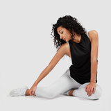 Woman in a black tank top and white leggings stretches gracefully on a plain background. Her expression is calm and focused, conveying tranquility.