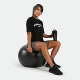  a fit woman sitting on an exercise ball holding a water bottle ,she wearing t-shirt black with short sage grey ,plus the shoes black 