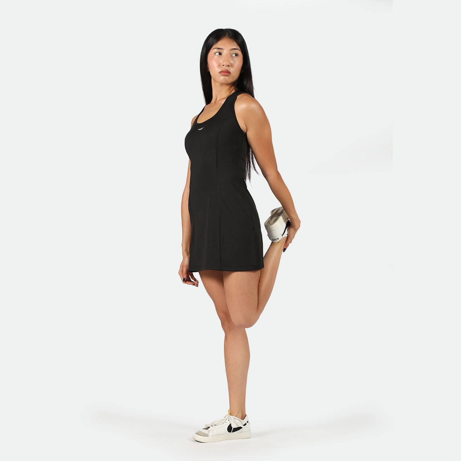 Woman in a black athletic dress stretching her leg behind her with one hand, standing on one foot. She wears white sneakers, exuding a focused demeanor.