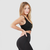 A fit woman with long blonde hair wearing a black sports bra and high-waisted black leggings. She is posing in a side profile with one hand on her hip, showcasing the fit and design of the activewear. The background is plain light gray