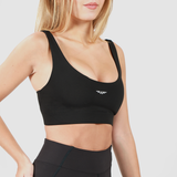 A fit woman with long blonde hair wearing a black sports bra and high-waisted black leggings. She is posing with one hand on her hip, highlighting the snug fit and design of the sports bra. The background is plain light gray.