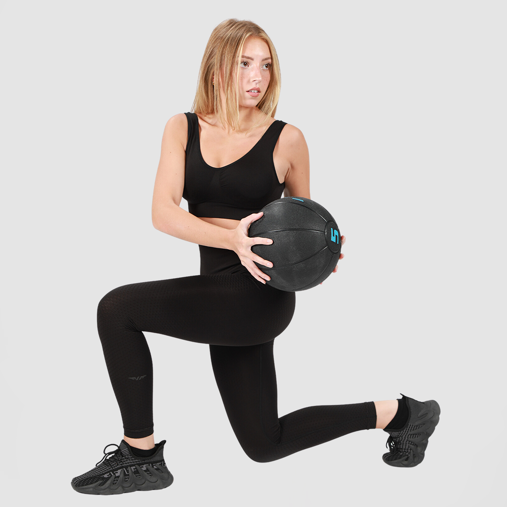 A young woman with shoulder-length blonde hair wearing a black sports bra, high-waisted leggings, and black sneakers, performing a lunge exercise while holding a black medicine ball, against a plain light gray background
