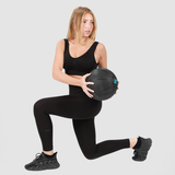A young woman with shoulder-length blonde hair wearing a black sports bra, high-waisted leggings, and black sneakers, performing a lunge exercise while holding a black medicine ball, against a plain light gray background