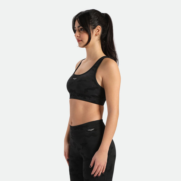 A woman in a camo sports bra and matching leggings stands in a three-quarter pose, the bra and legging have the same winneforce logo white In different directions, the background is white