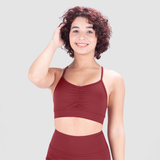 A young woman with short, curly hair smiles confidently while wearing a red sports bra and matching high-waisted leggings. She poses with one hand touching her hair against a plain light gray background.