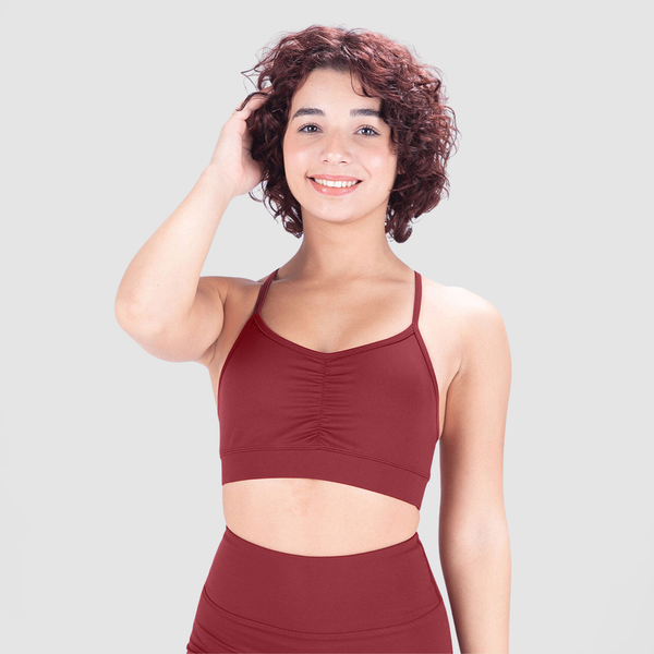 A young woman with short, curly hair smiles confidently while wearing a red sports bra and matching high-waisted leggings. She poses with one hand touching her hair against a plain light gray background.