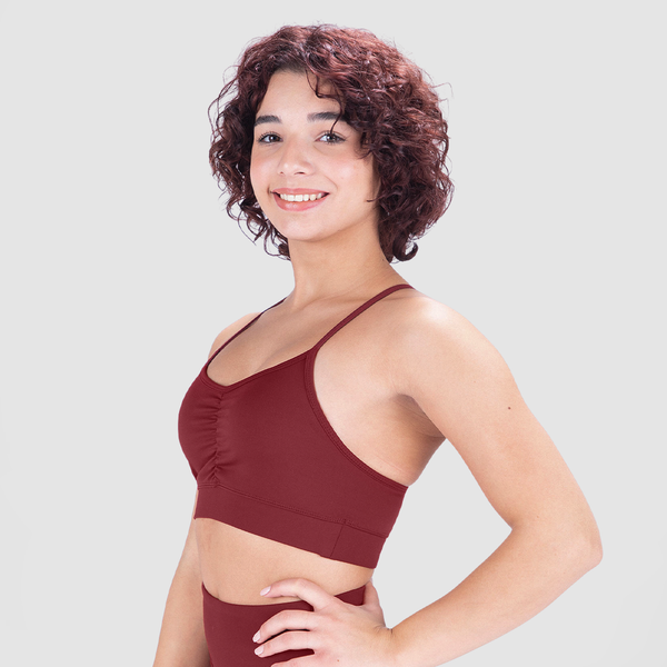 A young woman with short, curly hair smiles while posing in a burgundy sports bra and matching high-waisted leggings. She stands in a three-quarter profile with one hand on her hip against a plain light gray background
