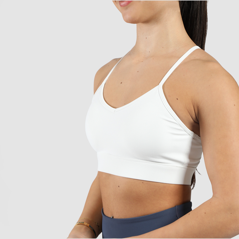 A woman wearing a white sports bra and navy high-waisted leggings, smiling slightly against a neutral gray background.