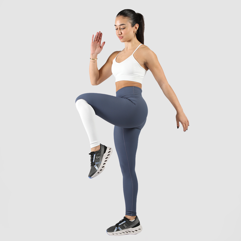 A woman wearing a white sports bra and dark blue leggings stands to the side, displaying fitness-focused clothing against a plain background.