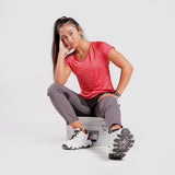 A woman in athletic wear sits on a silver metal case, resting her head on one hand. She wears a red short-sleeve top, gray pants, and white sneakers with star-patterned soles,