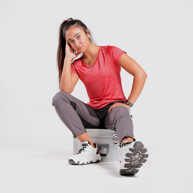 A woman in athletic wear sits on a silver metal case, resting her head on one hand. She wears a red short-sleeve top, gray pants, and white sneakers with star-patterned soles,