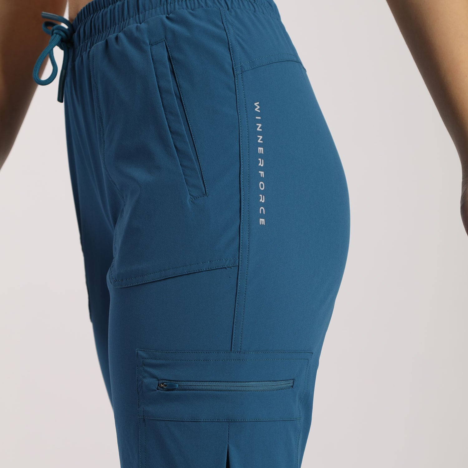  A close-up view of a person wearing blue cargo jogger pants with multiple pockets, including a zippered pocket on the thigh and a vertical printed text design on the side. The pants have an elastic waistband with a drawstring