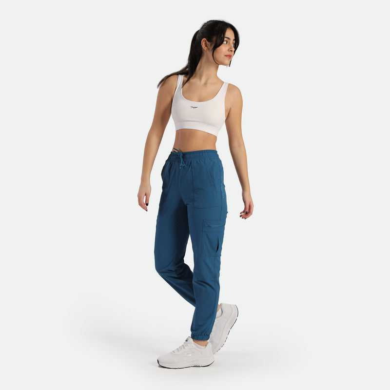  A woman wearing blue cargo jogger pants paired with a white sports bra and white sneakers. She stands in a relaxed pose against a light background, showcasing the athletic and casual fit of the joggers.
