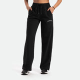 a fit woman wearing pants with high waisted ,plus white logo in front, she wearing black shoes athletic ,the background is light grey 
