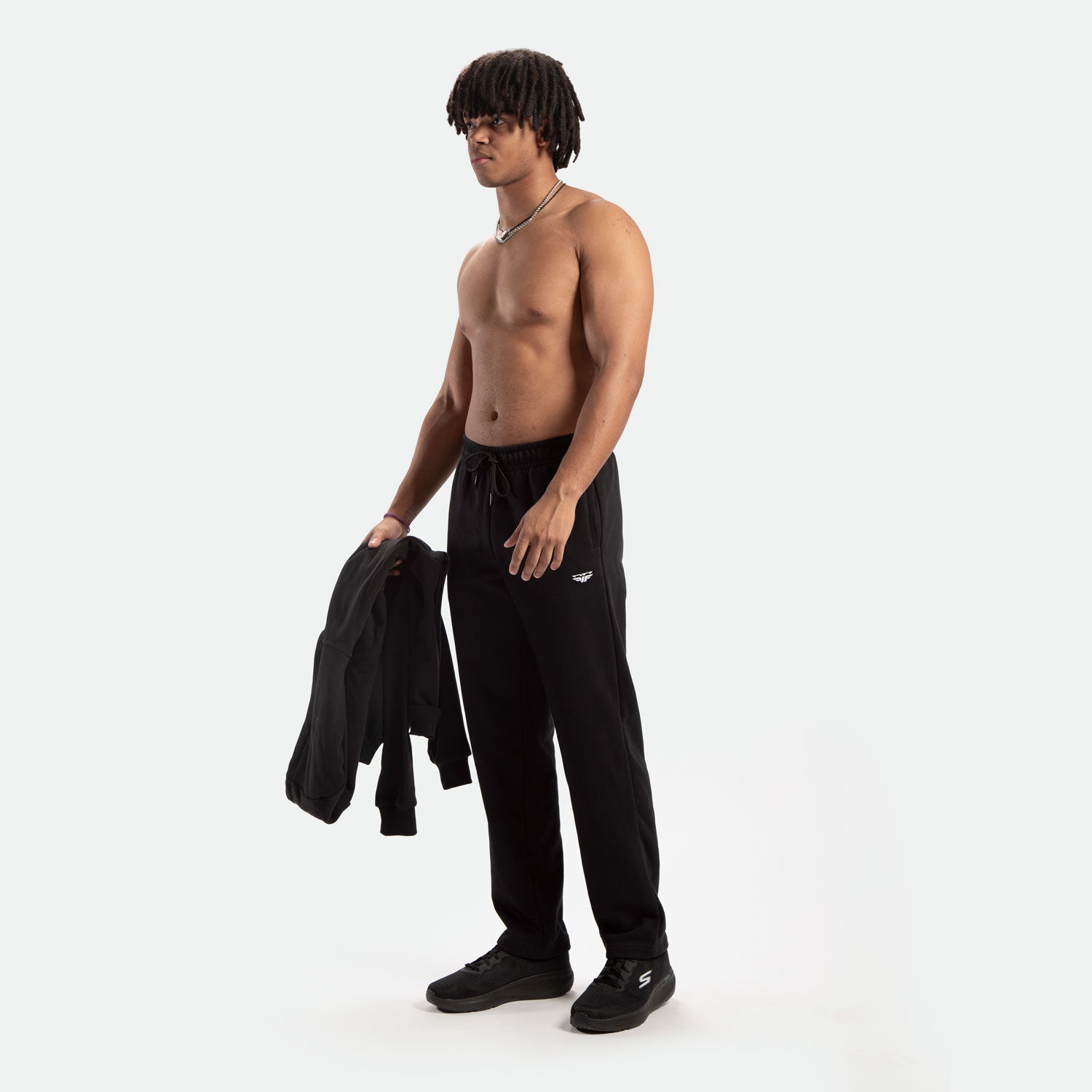 Shirtless man with dreadlocks stands confidently, holding a black jacket. He's wearing black sweatpants and sneakers, set against a white background.