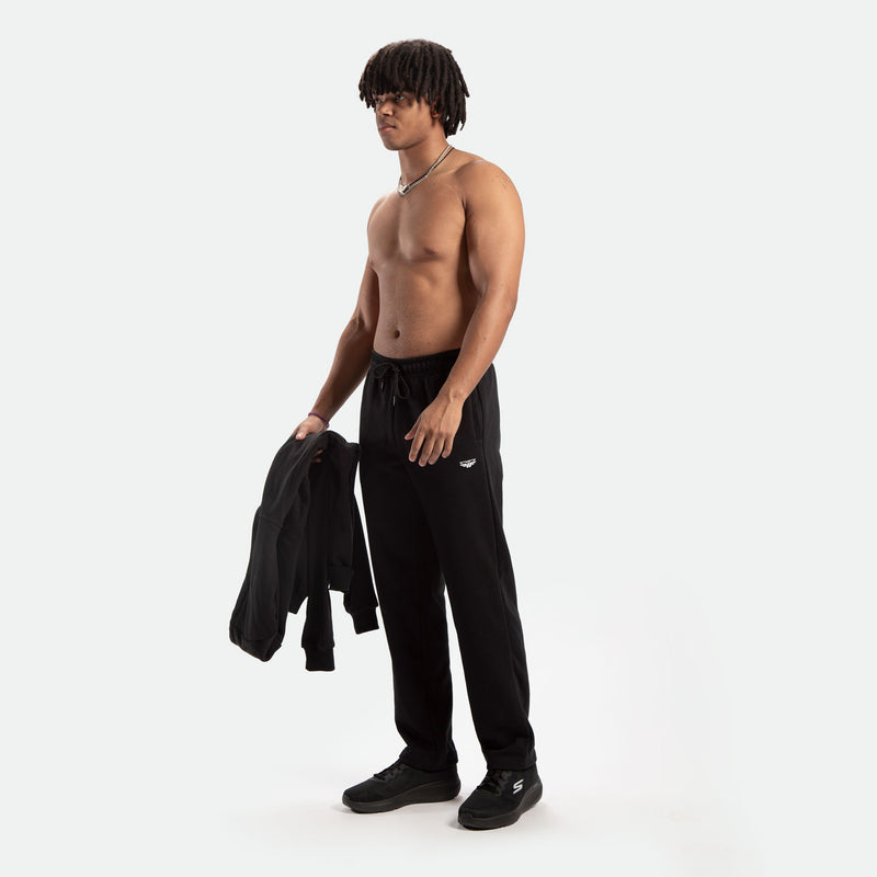 Shirtless man with dreadlocks stands confidently, holding a black jacket. He's wearing black sweatpants and sneakers, set against a white background.