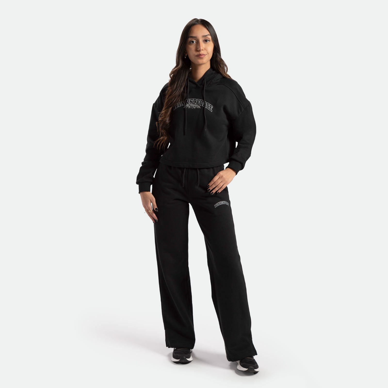 a fit woman with long hair brown look at the camera and smile , she wearing matching hoodie and pants black with logo, she wearing black shoes athletic, plus the background is light grey