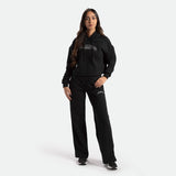 a fit woman with long hair brown look at the camera and smile , she wearing matching hoodie and pants black with logo, she wearing black shoes athletic, plus the background is light grey
