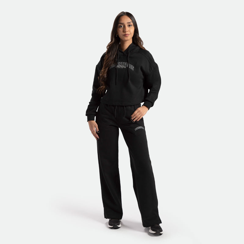a fit woman with long hair brown look at the camera and smile , she wearing matching hoodie and pants black with logo, she wearing black shoes athletic, plus the background is light grey