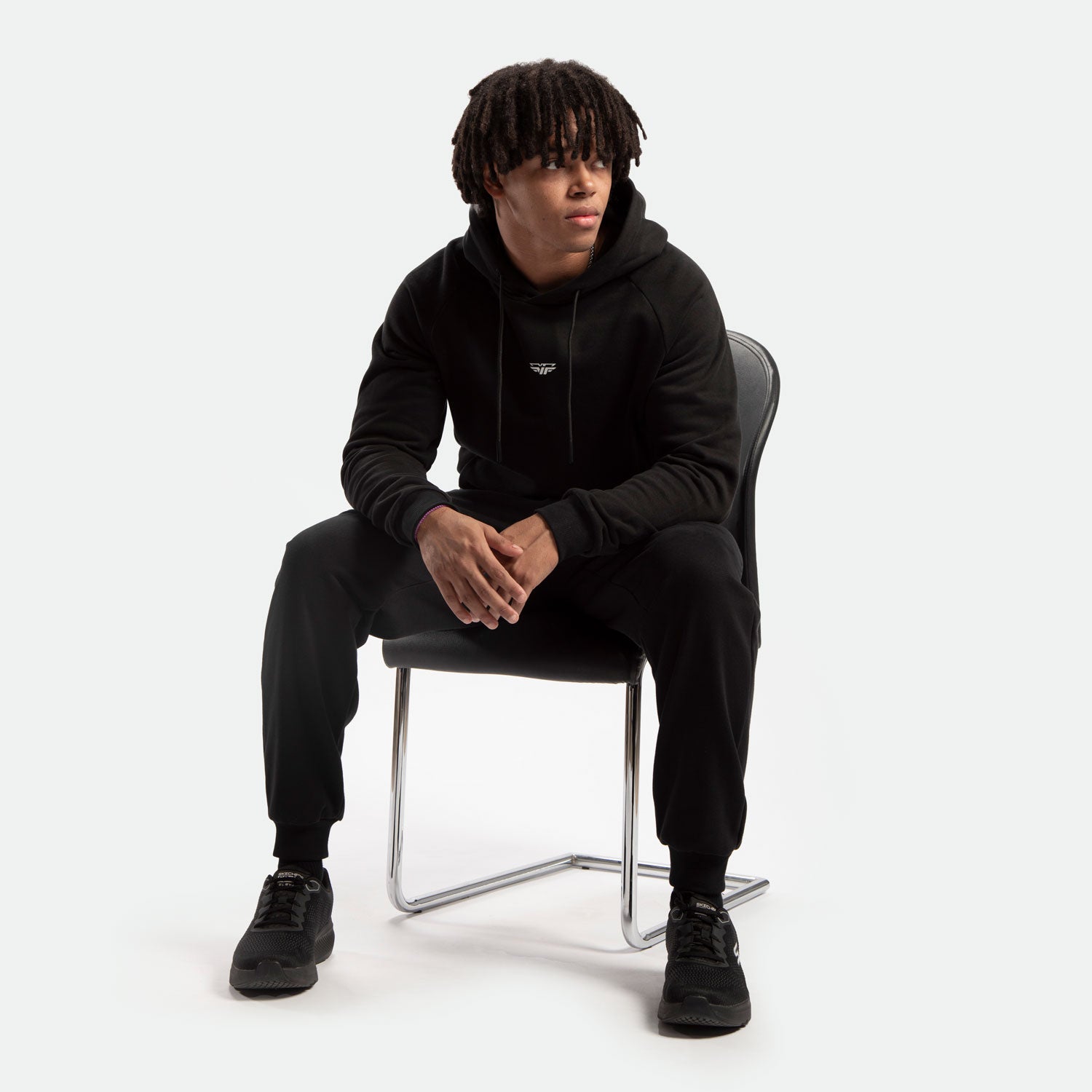 A person with short dreadlocks sits on a chair, wearing a black hoodie and matching pants. They appear relaxed and contemplative, looking to the side.