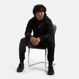 A person with short dreadlocks sits on a chair, wearing a black hoodie and matching pants. They appear relaxed and contemplative, looking to the side.